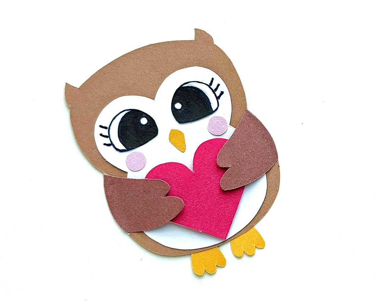 Cute brown paper owl with white front, black eyes, and pink cheeks, holding a red heart