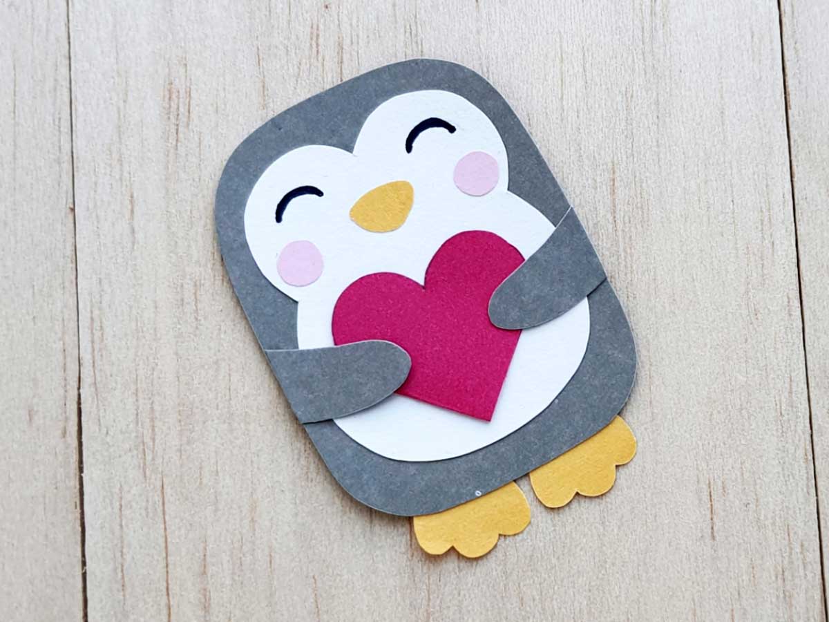 Image of gray and white paper penguin holding a red paper heart on a light wood background