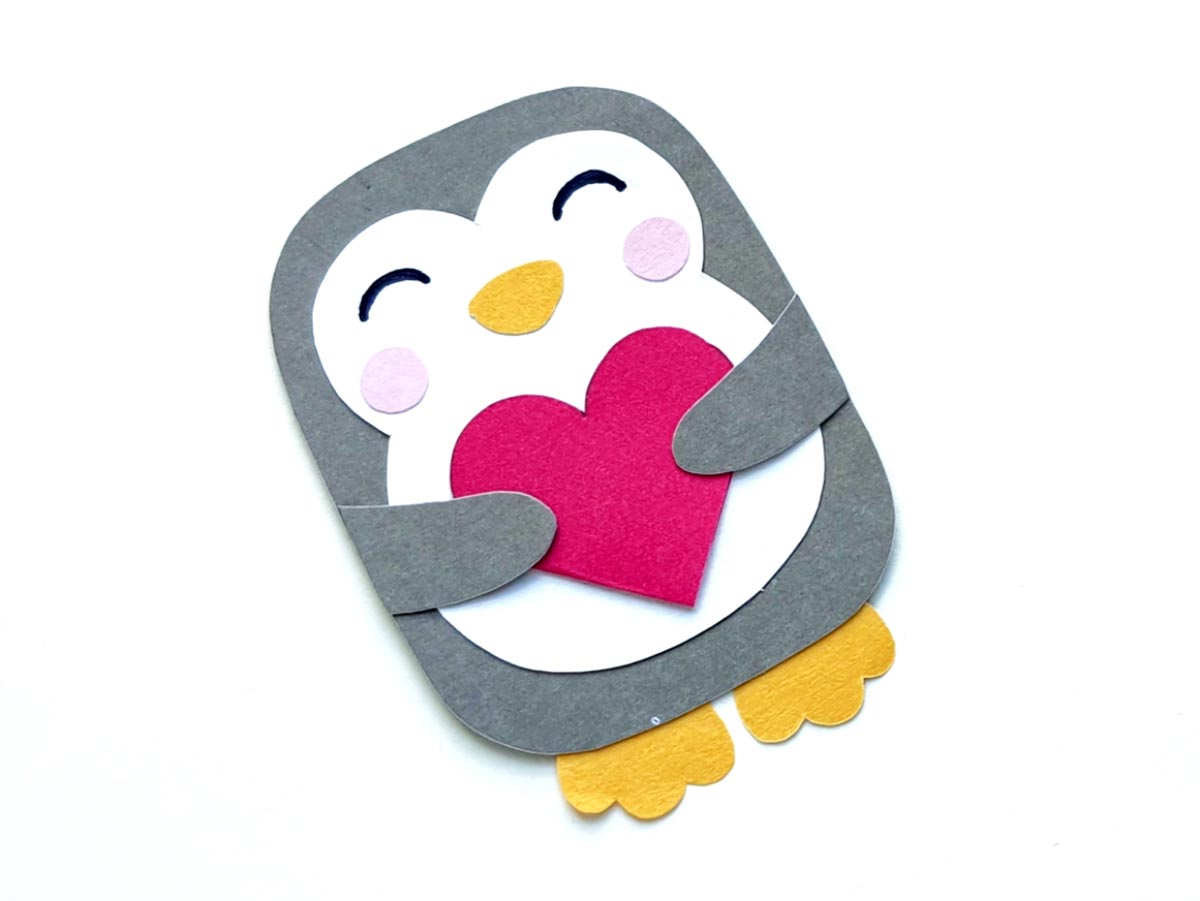Image of gray and white paper penguin holding a red paper heart on a white background