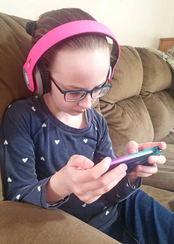 Image of school girl wearing a blue shirt with white hearts, with blue glasses and pink headphones, holding a phone