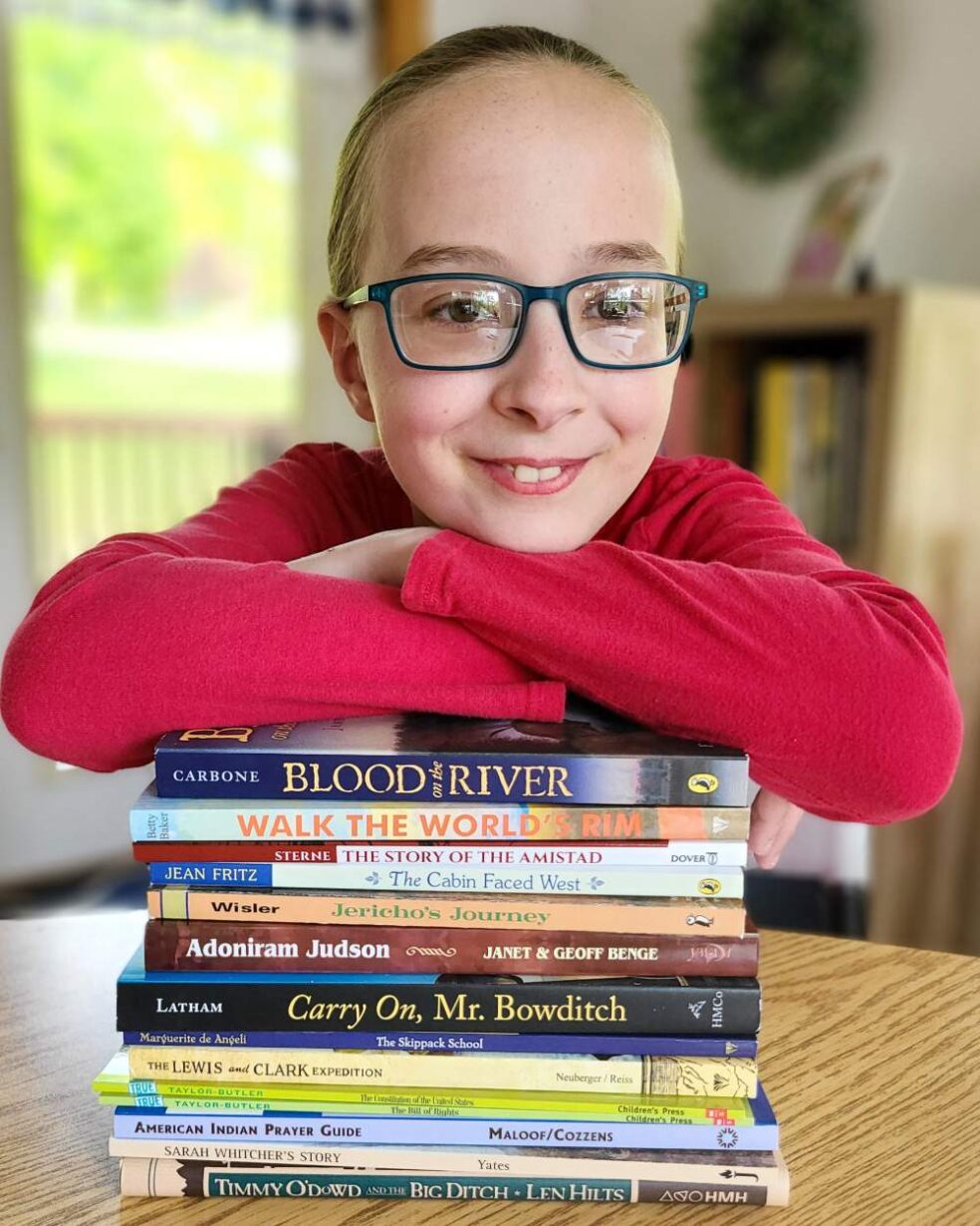 Smiling girl in red shirt with blue glasses, leaning her arms on a stack of living history books