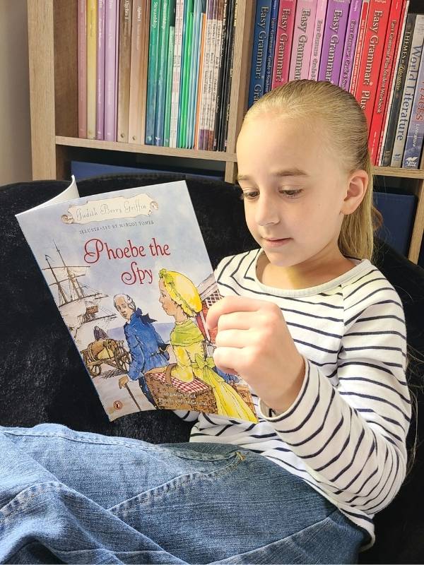Image of young girl in striped shirt sitting in front of bookcase and learning through literature entitled Phoebe the Spy