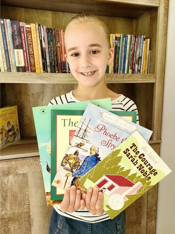 Smiling young girl standing in front of bookcase holding a selection of history books for learning through literature