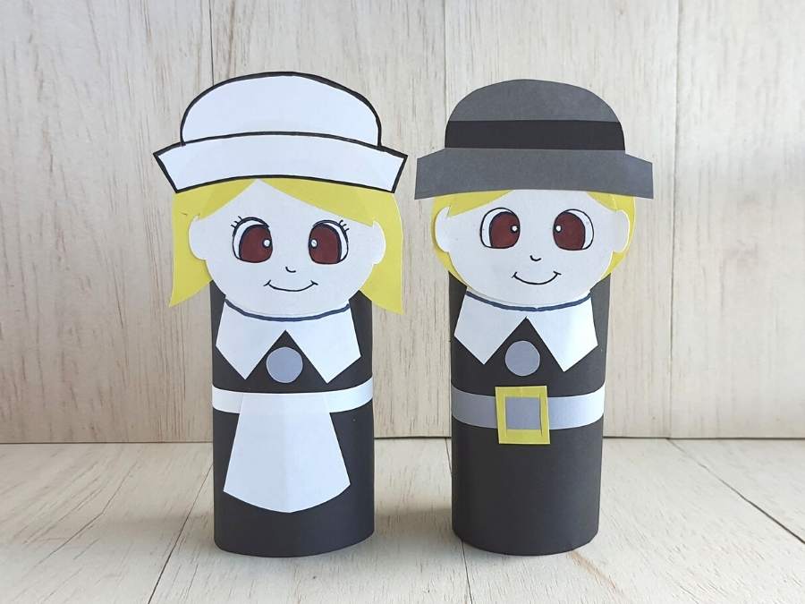 Image of girl and boy Pilgrims constructed from empty toilet paper rolls and construction paper in front of a light gray wooden background.
