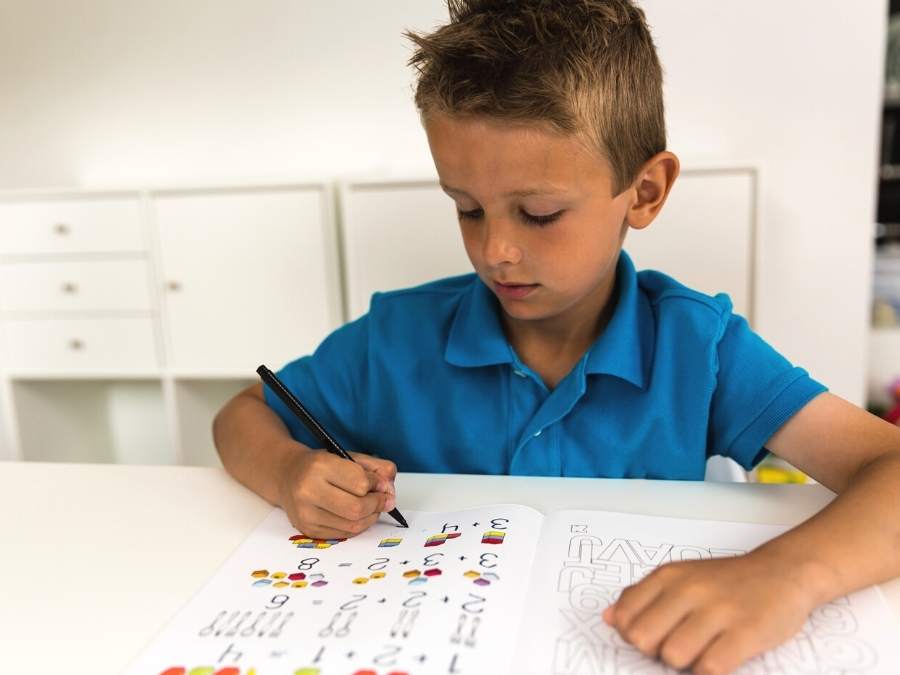 Image of young boy in blue shirt sitting at a table and working in a math workbook to catch up math
