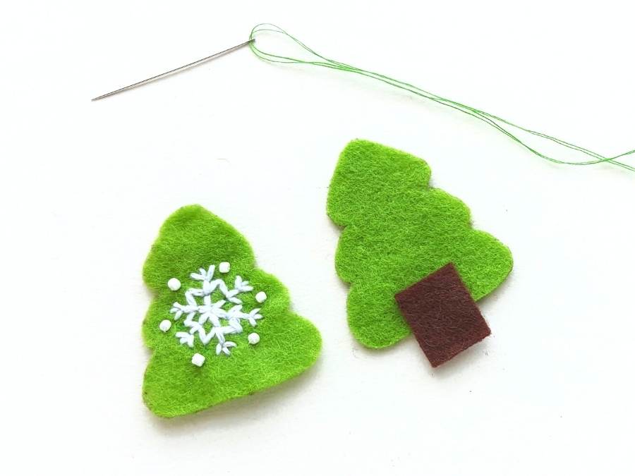 Image of 2 green felt Christmas tree cutouts, one with a snowflake embroidered in white on the front, and the other with a piece of brown felt attached for a trunk.