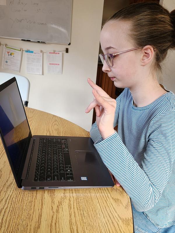 Image of young girl signing along with an online ASL class on a laptop.