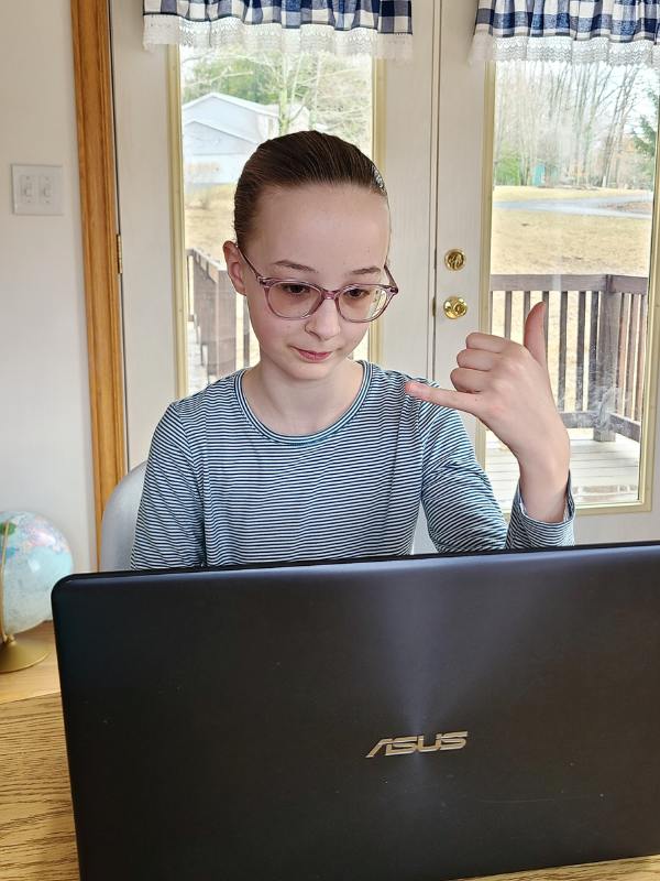 Image of young girl in blue-striped shirt, sitting at a laptop and practicing ASL.