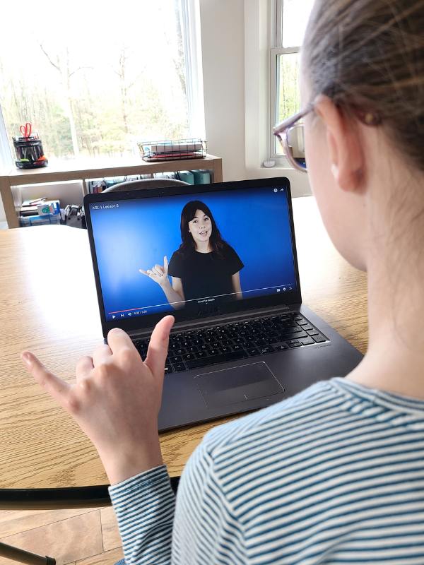 Image of young girl in blue striped shirt practicing ASL along with an online instructor.