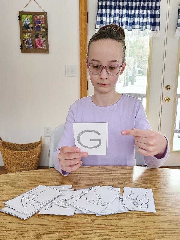 Image of girl in purple shirt with pink glasses holding a flash card with the letter and G and finger spelling it in ASL.