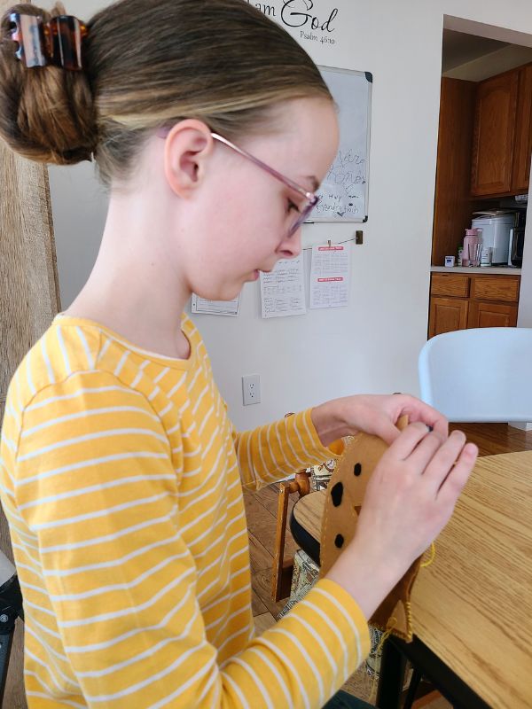 Image of girl in yellow striped shirt sitting at a table and sewing a brown felt fox stuffie.