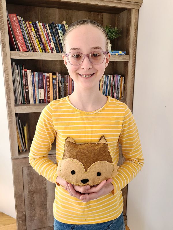 Image of smiling girl in yellow striped shirt standing in front of a bookcase and holding a brown felt fox stuffie.