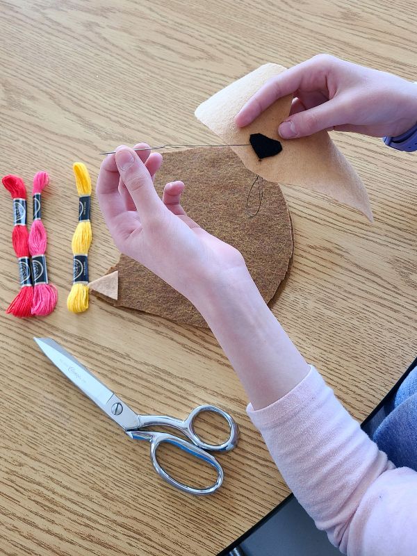 Image of girl's hands sewing a black nose onto a tan felt face, with colorful embroidery floss and scissors in the background.