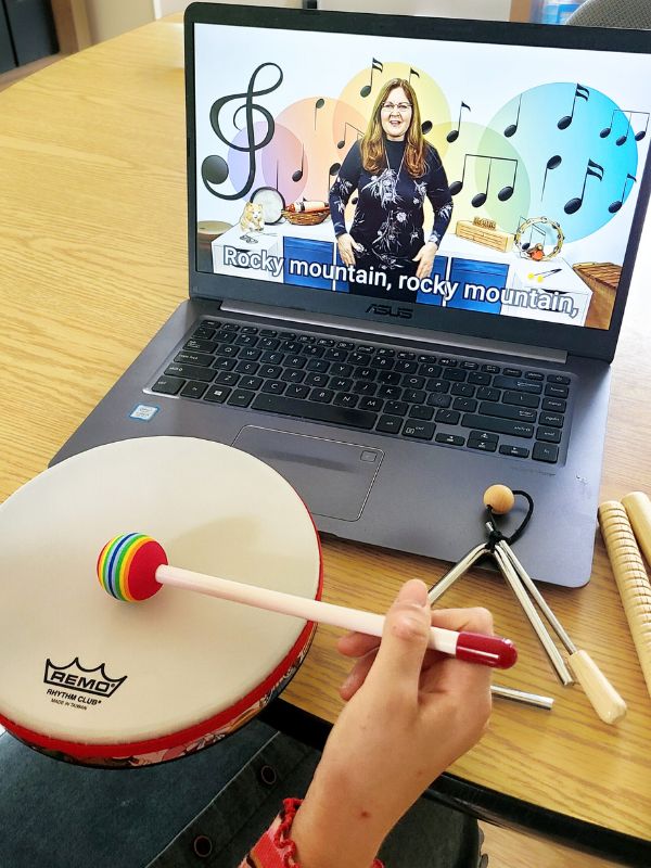 Online music lessons Image of laptop displaying an online music lesson in the background, with a girl's hand using a soft drumstick to demonstrate rhythm on a drum.