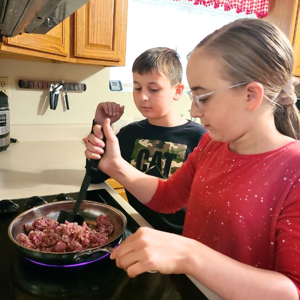 Online cooking lessons for kids Image of tween girl cooking ground beef in a skillet while a younger boy watches.