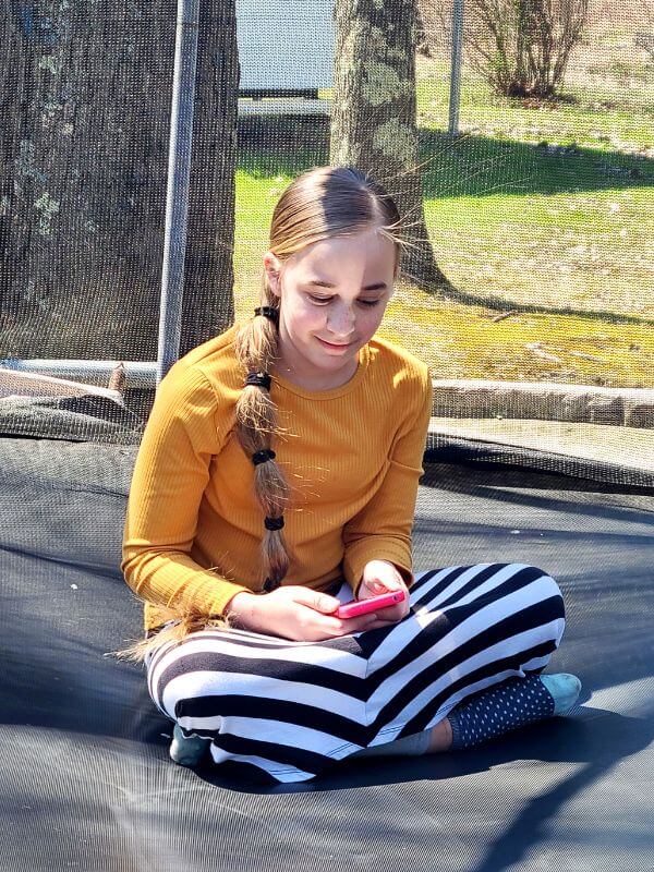 Homeschool country studies Image of young girl sitting on trampoline, holding her phone and listening to Stories Around the World.