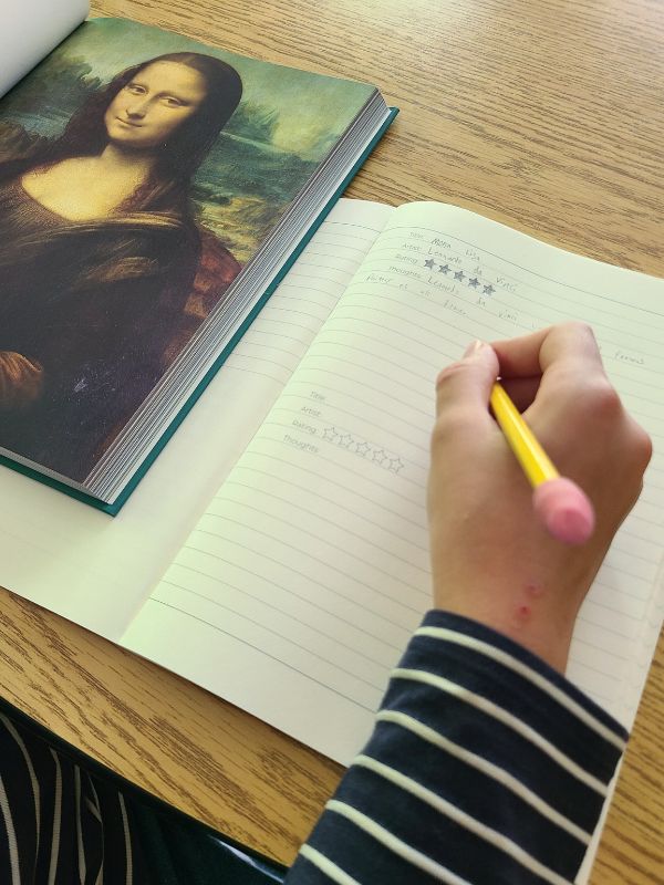 Charlotte Mason picture study Image of young girl's hand writing in a notebook with an open art book beside her.