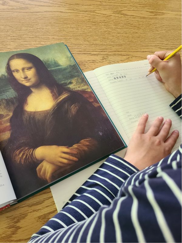 Homeschool fine arts elective Image of girl writing in a notebook while studying a book of artists.